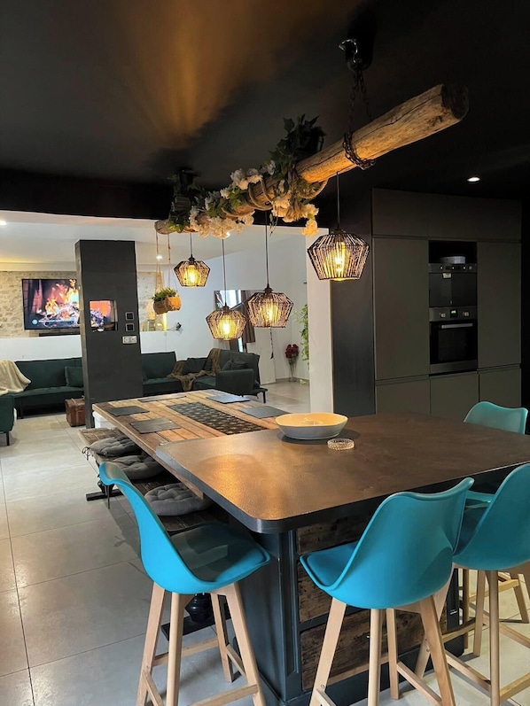 Una cocina moderna con una isla central, taburetes de bar azules, luces colgantes y una pared de piedra con chimenea.