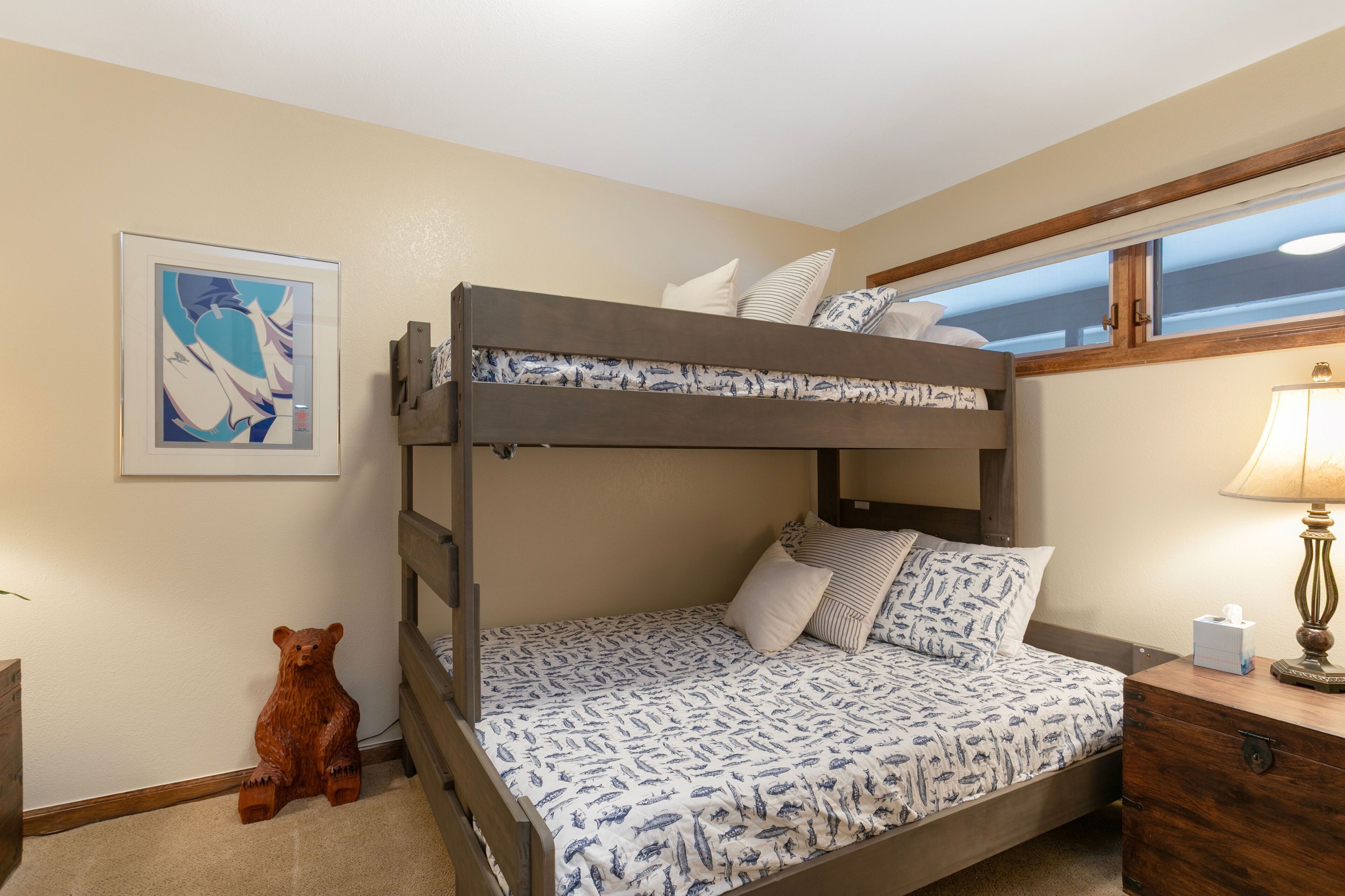 A bunk bed with a patterned bedspread, a bedside table with a lamp, a wooden bear decoration, and a framed picture on the wall.