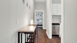 A hallway leading to a bedroom with a bed, a bedside table, and a nightstand. There is a small table with a lamp in the foreground.