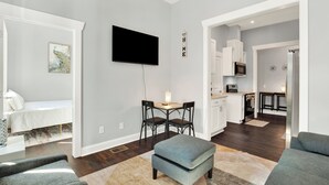 A modern living room with a flat-screen TV, a dining area with a table and chairs, a kitchenette, and a grey sofa.