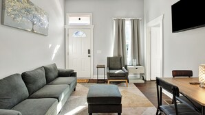 A modern living room with a grey sofa, a wooden dining table with chairs, a grey armchair, and a flat-screen TV mounted on the wall.