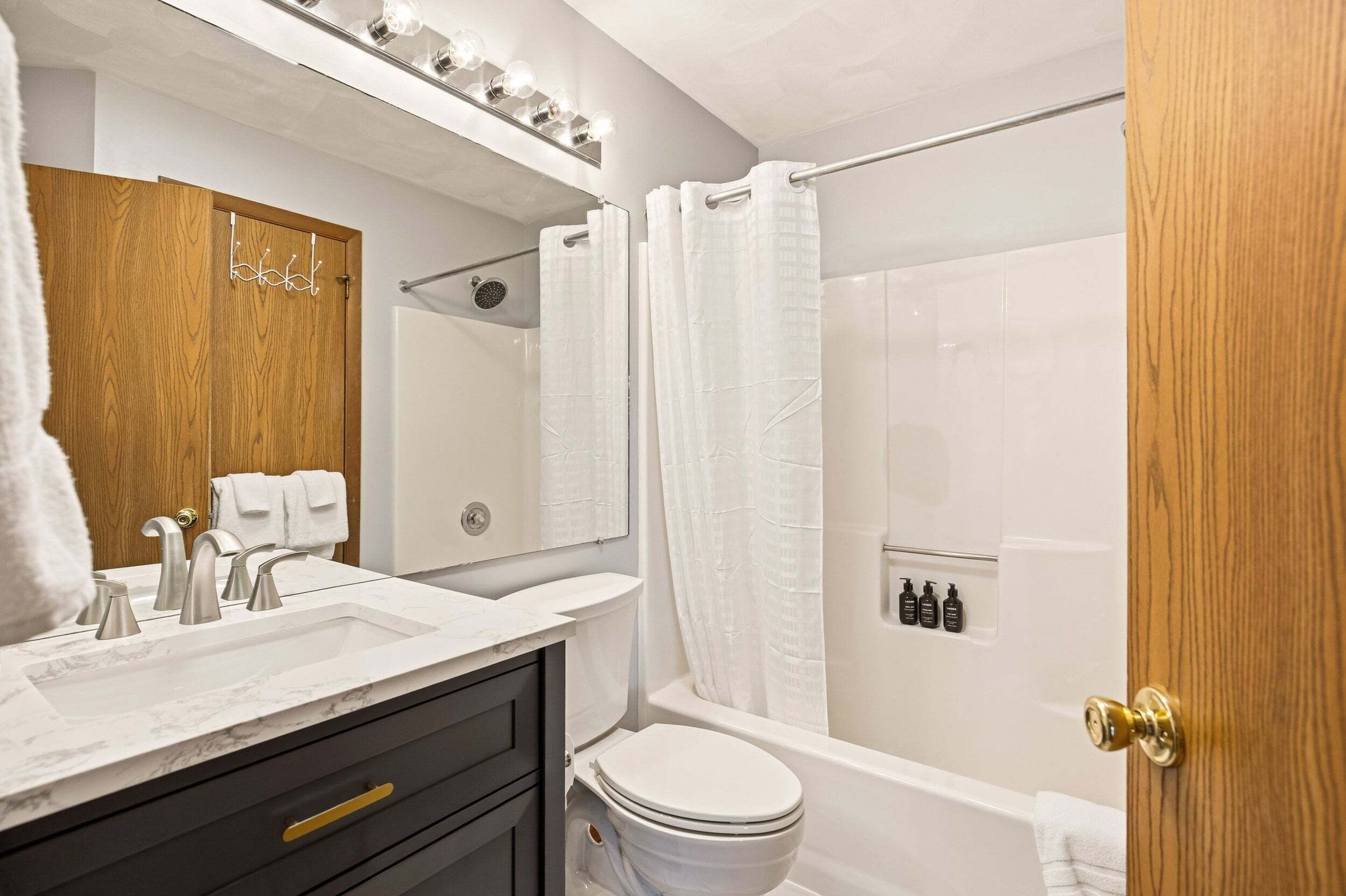 A modern bathroom with a marble sink, a wooden vanity, a white toilet, a bathtub with a shower curtain, and a mirror with lights above.