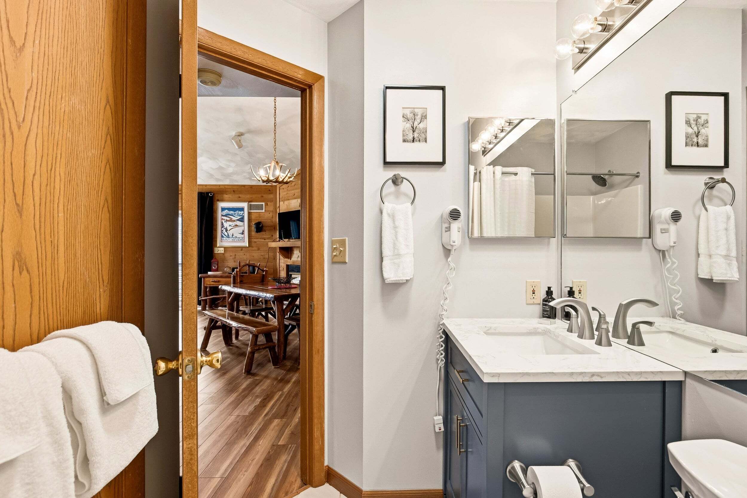 A bathroom with dual sinks, a large mirror, and a wooden door leading to a dining area.