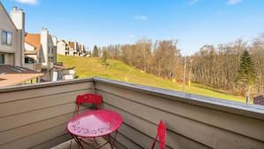 Un balcón con una mesa y sillas rojas, con vista a un área verde con árboles y casas al fondo.