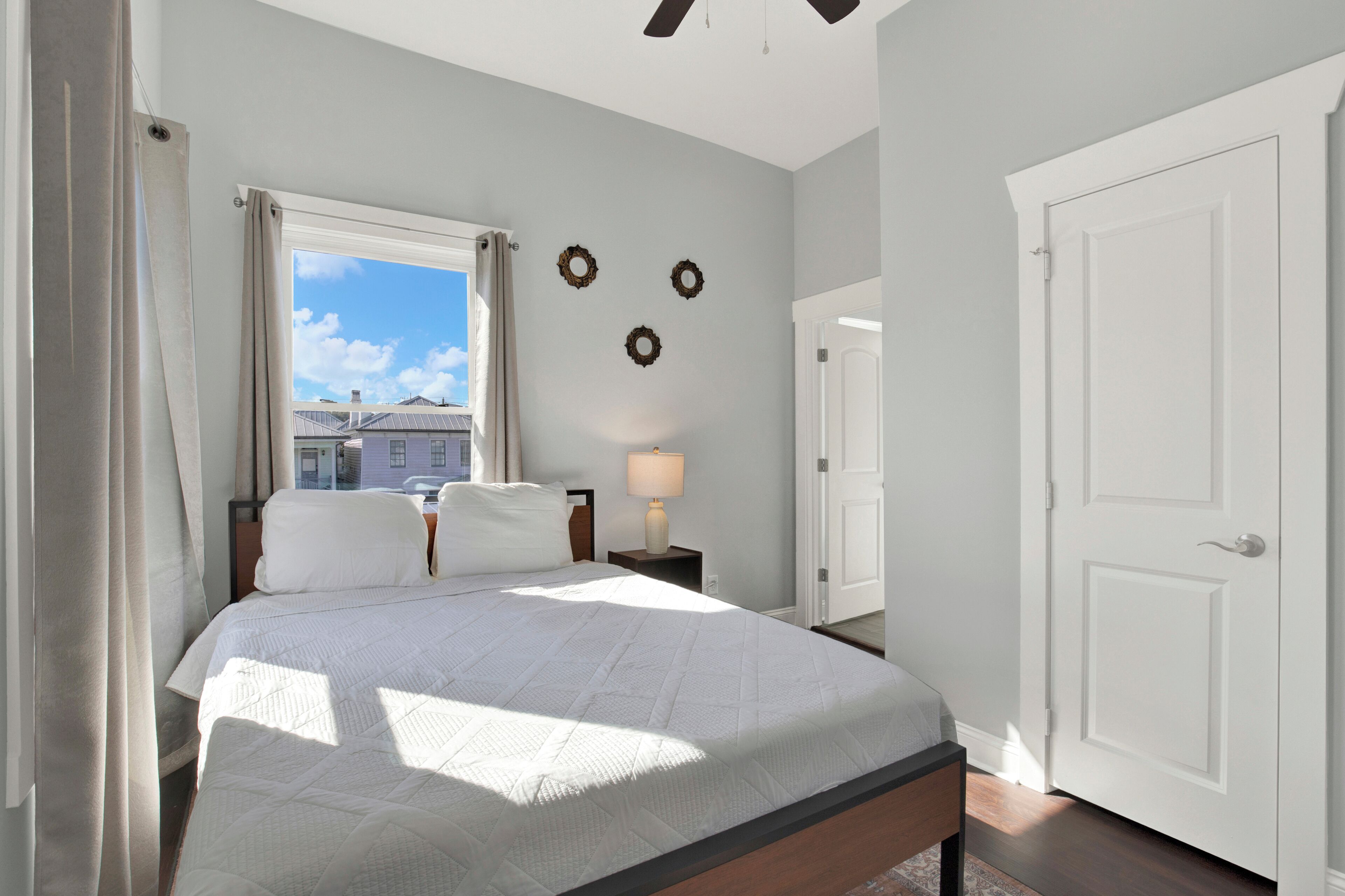 Un dormitorio con cama, una mesita de noche con lámpara, una ventana con cortinas y una puerta.