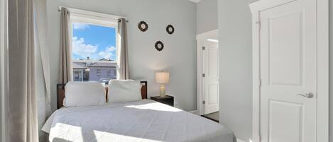 Un dormitorio con cama, una mesita de noche con lámpara, una ventana con cortinas y una puerta.