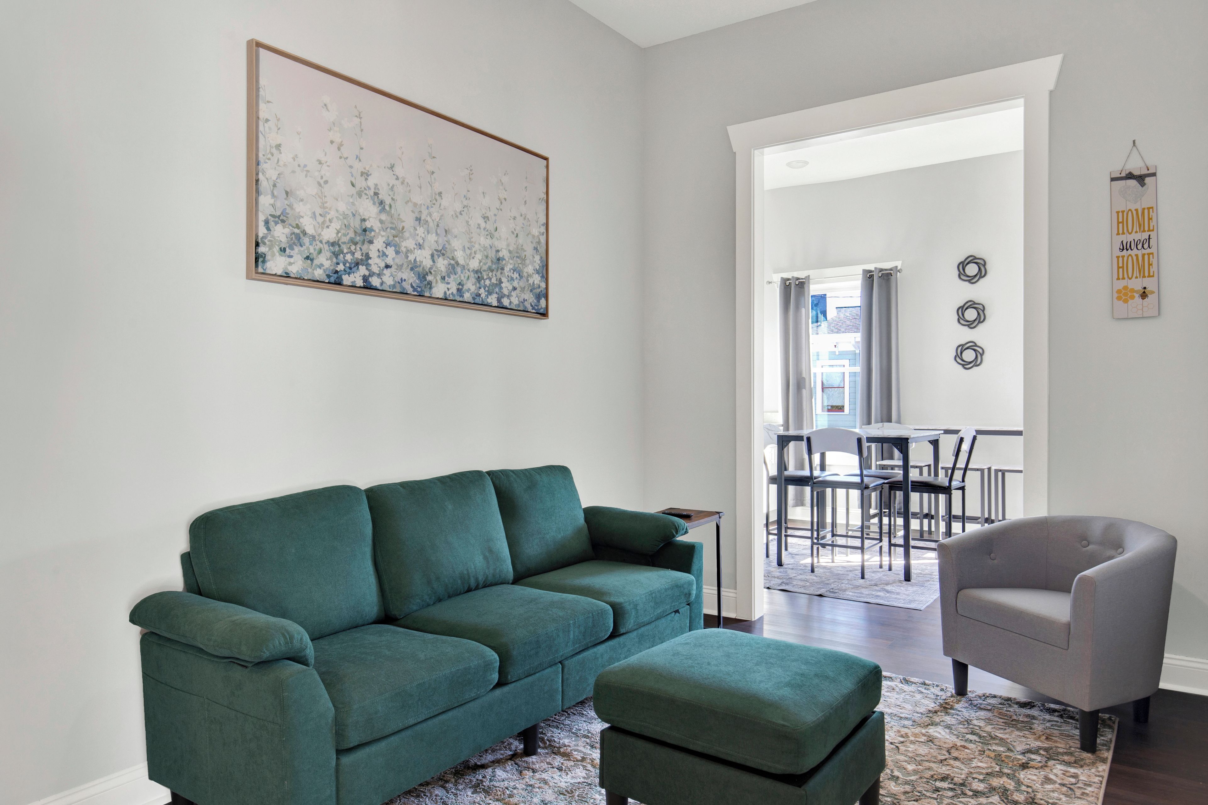Een woonkamer met een groene bank, een grijze fauteuil en een bijpassend voetenbankje. Aan de muur hangt een schilderij en een bord met de tekst “HOME sweet HOME”.
