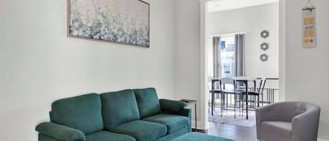 A living room with a green sofa, a grey armchair, and a matching ottoman. There is a painting on the wall and a sign that reads "HOME sweet HOME".