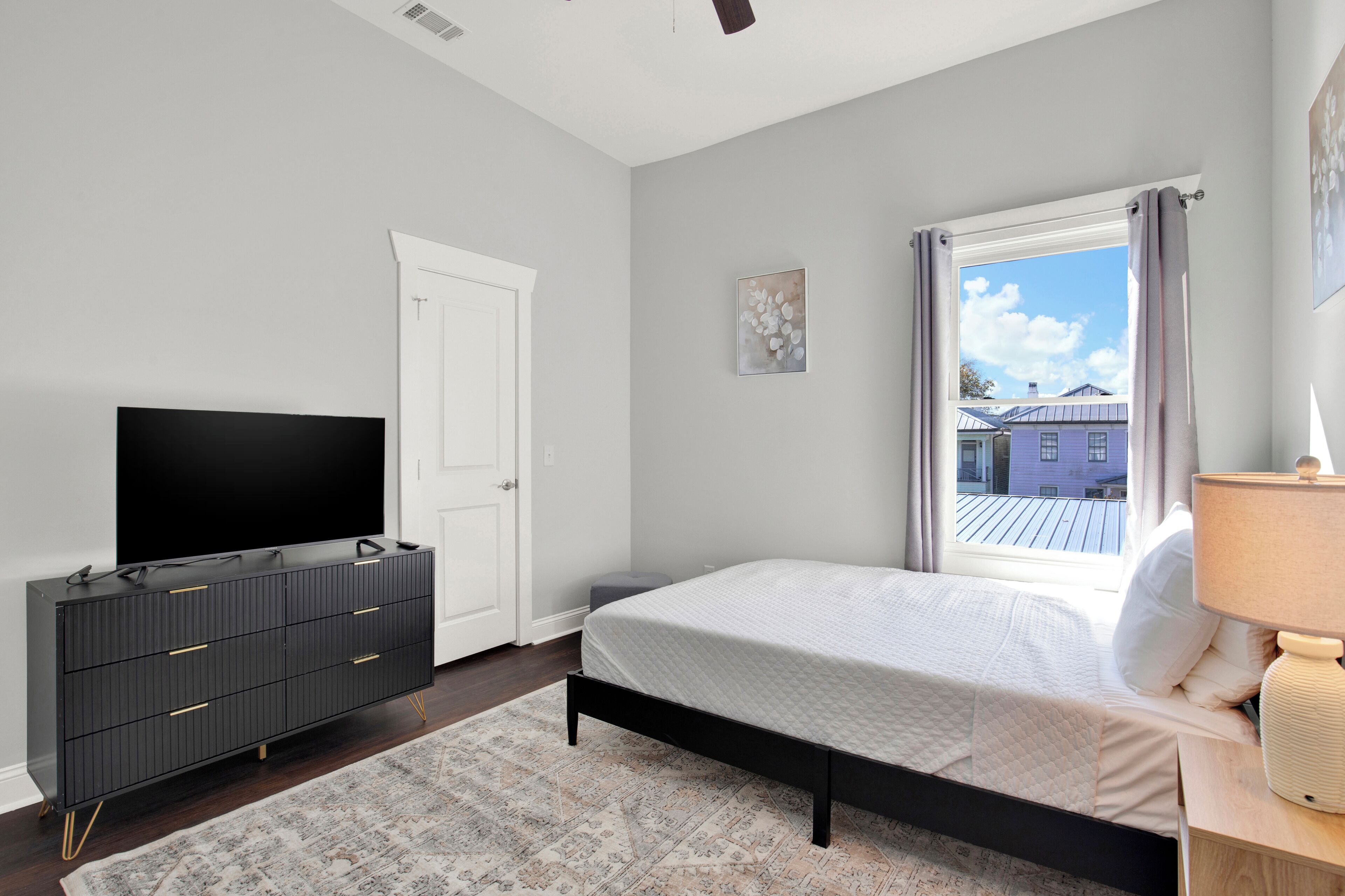 Une chambre à coucher avec un lit, une télévision, une commode, une fenêtre avec des rideaux et une vue sur les bâtiments à l’extérieur.