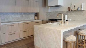A modern kitchen with light wood cabinets, a marble countertop, a gas stove, and a sink with a gold faucet. There are two stools with cushions at the island. - 1175 B Casolar Del Norte 3 Bedroom Duplex (Vail)