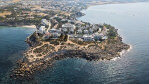 An aerial view of a coastal town with a rocky shoreline, clear blue waters, and a cluster of white buildings with a central road. - Lantana Villas and Hotel (Pegeia)