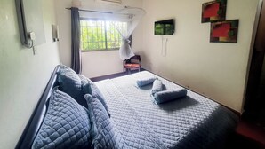 A bedroom with a bed, a window with curtains, a bedside table, and a wall-mounted TV. - Acamms Lodge (Lusaka)