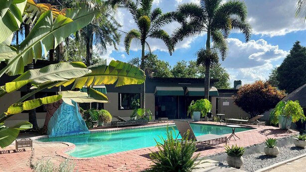 A swimming pool with a waterfall feature, surrounded by palm trees and potted plants. - Acamms Lodge (Lusaka)