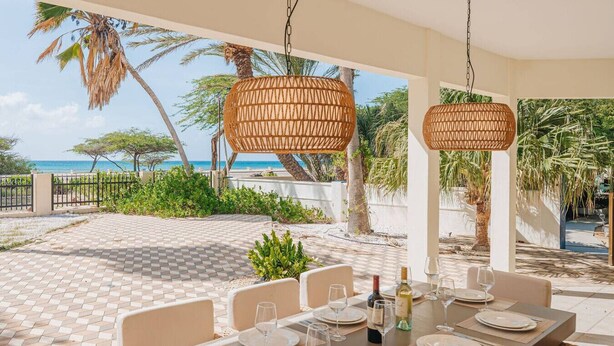 An outdoor dining area with a view of the sea, featuring a table set for six with plates, glasses, and a bottle of wine. - NEW: Oceanfront Villa w/Private HotTub and Pool (Noord)