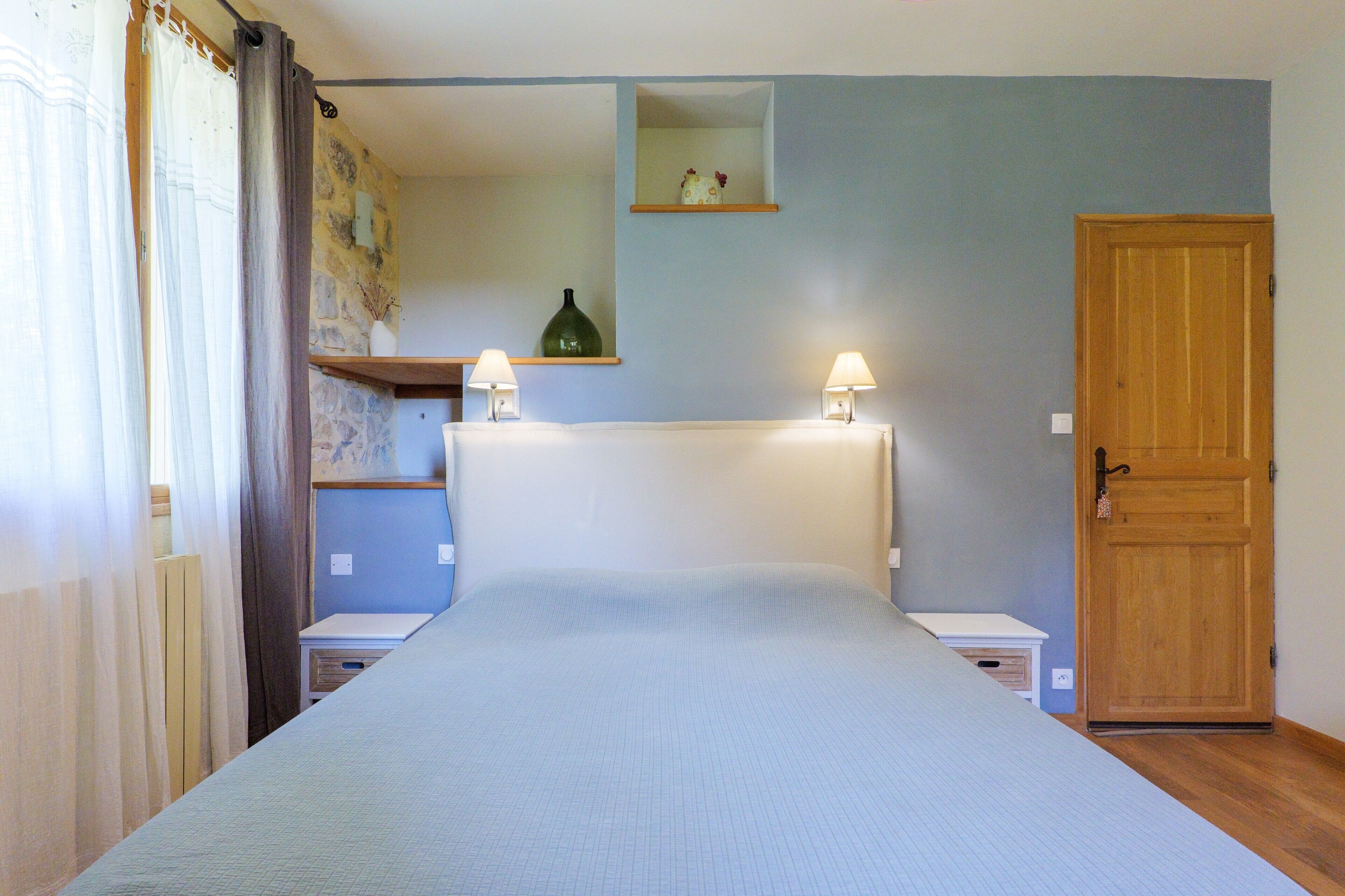Quarto com cama, duas luminárias de cabeceira, uma porta de madeira e uma pequena estante com um vaso.