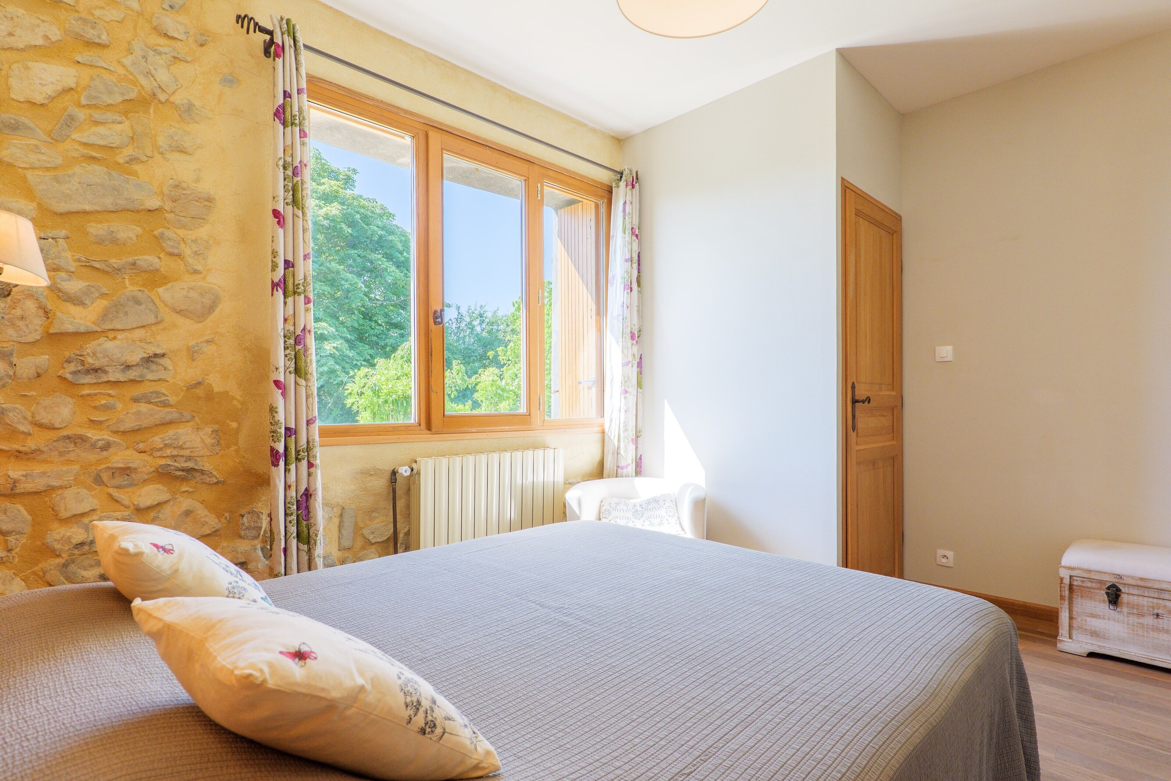 Une chambre avec un mur en pierre, un lit avec des oreillers, une fenêtre avec des rideaux, un radiateur et une porte en bois.