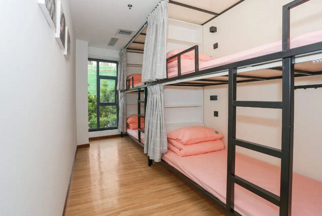 A dormitory room with bunk beds, a window with a view of greenery, and a wooden floor.