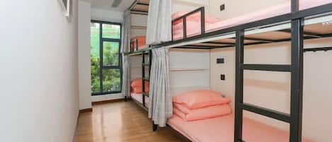 A dormitory room with bunk beds, a window with a view of greenery, and a wooden floor.