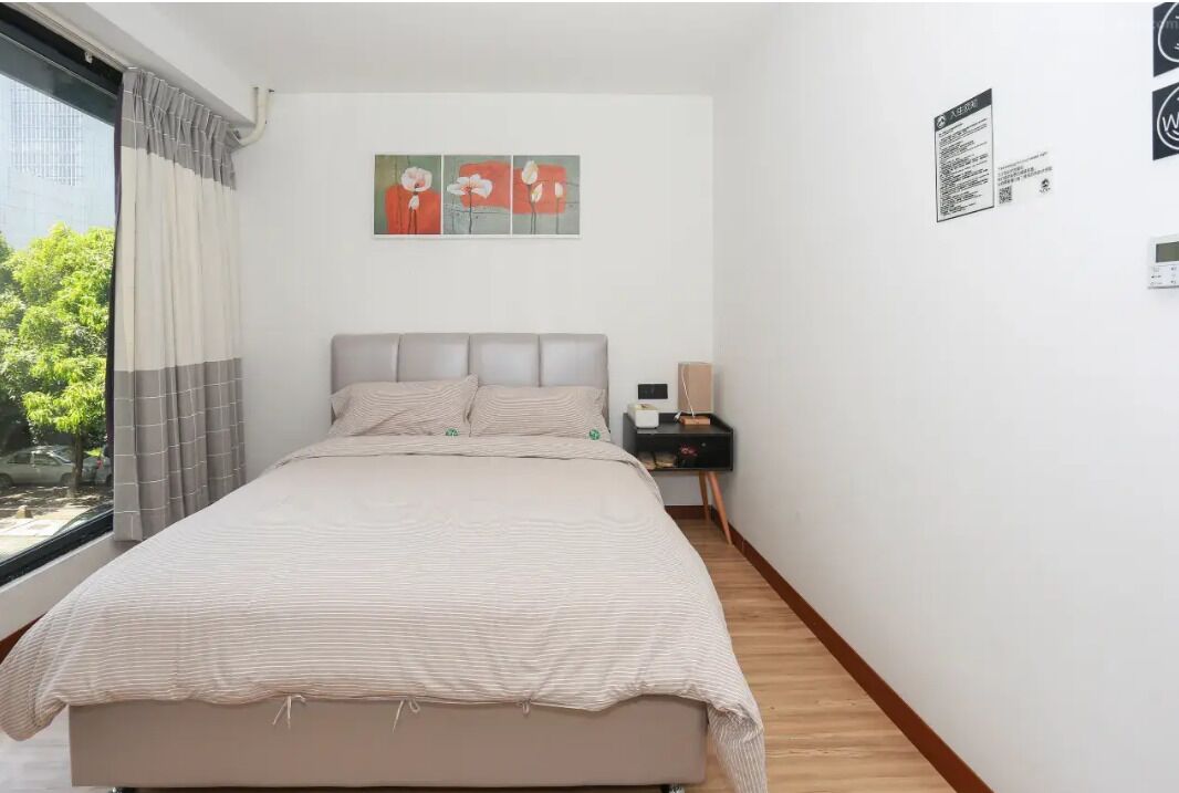 Quarto com cama, mesa de cabeceira, janela com cortinas e uma obra de arte na parede.