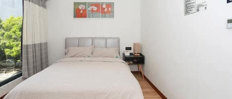 Quarto com cama, mesa de cabeceira, janela com cortinas e uma obra de arte na parede.