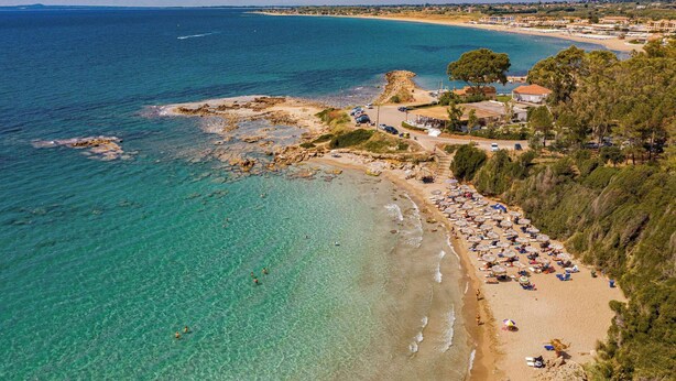 Vue aérienne d’une zone côtière avec une eau turquoise cristalline, une plage de sable et une rangée de parasols de plage.