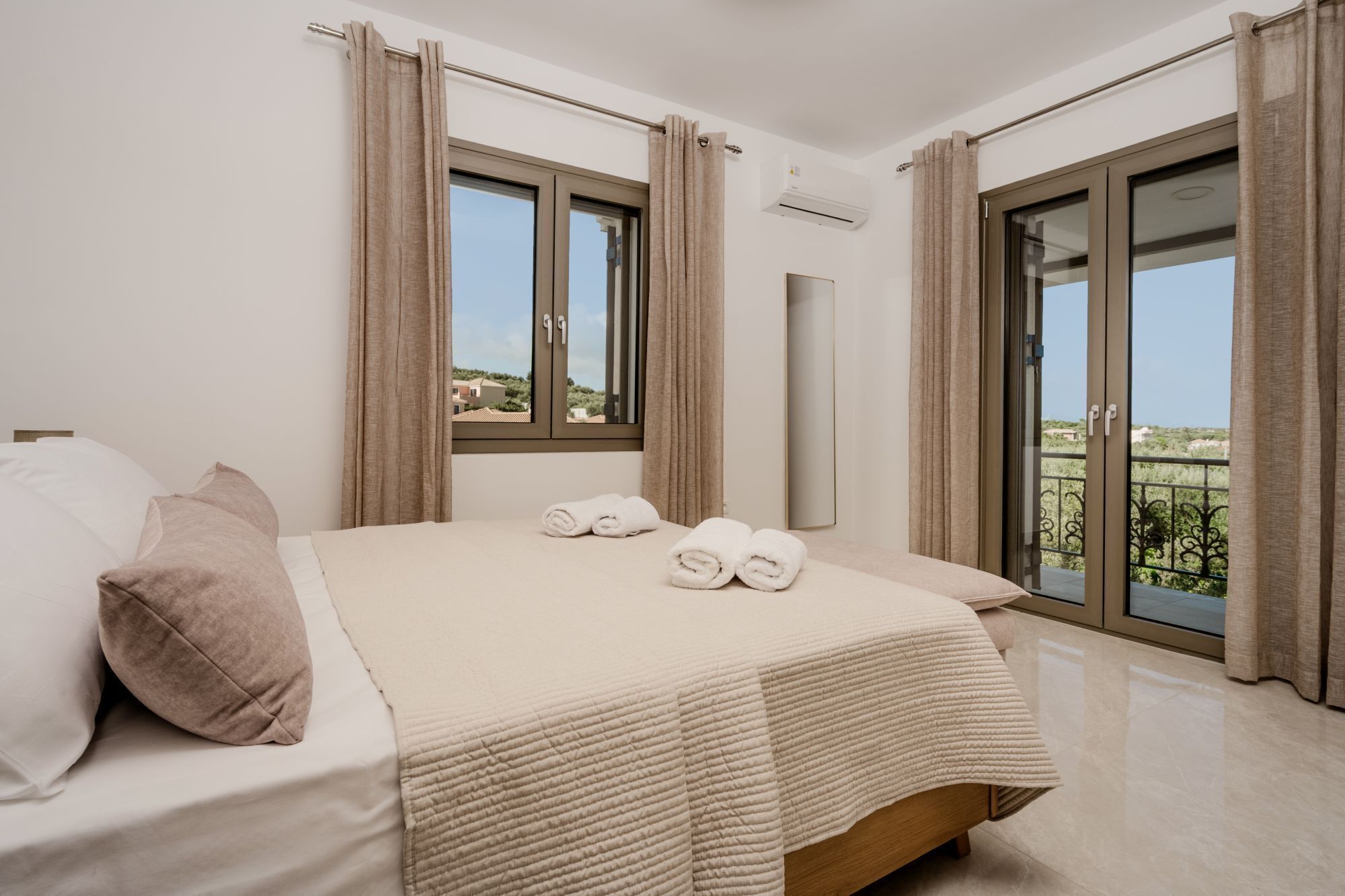A bedroom with a large bed, two windows, a balcony, and an air conditioning unit.