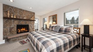A bedroom with a stone fireplace, a large bed with a plaid bedspread, two bedside lamps, and a view of the outdoors through a window. - Hi Country Haus Unit 2312 New, renovated 2 bedroom, private deck extra amenities (Winter Park)
