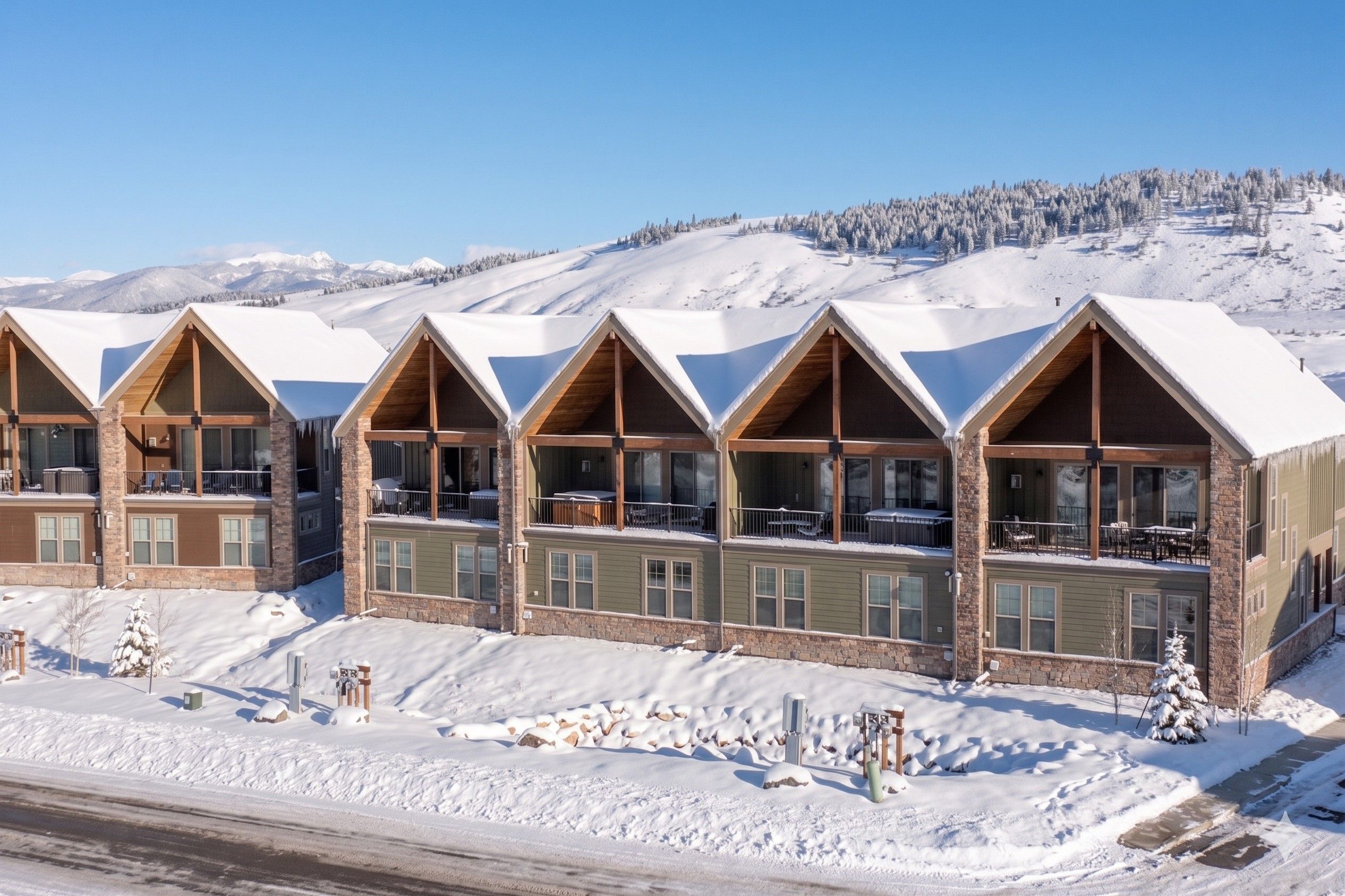 A row of chalet-style buildings with large windows and balconies, set against a snowy mountain backdrop.