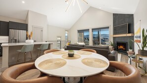 Eine moderne Küche mit einer zentralen Kochinsel, ein Essbereich mit rundem Tisch und ein Wohnzimmer mit Kamin.