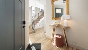 Ein Flur mit Holzboden, einer grauen Tür, einer Treppe, einem Spiegel, einem kleinen Tisch mit Lampe und einem roten Hocker.