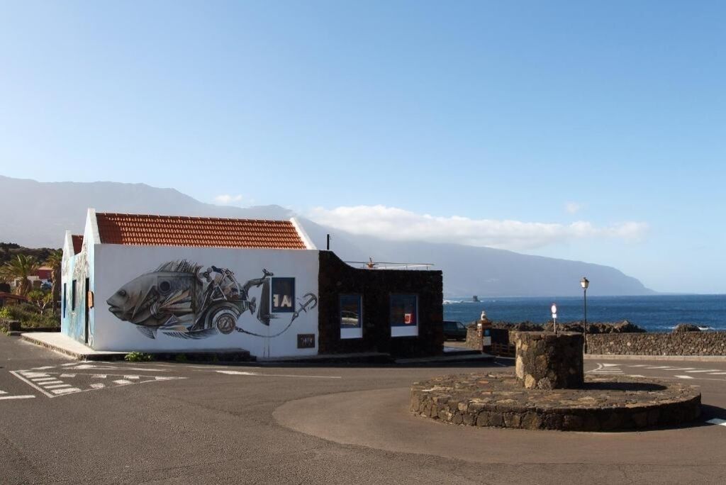 A building with a mural of a fish and a bicycle, a stone structure, and a view of the sea and mountains.