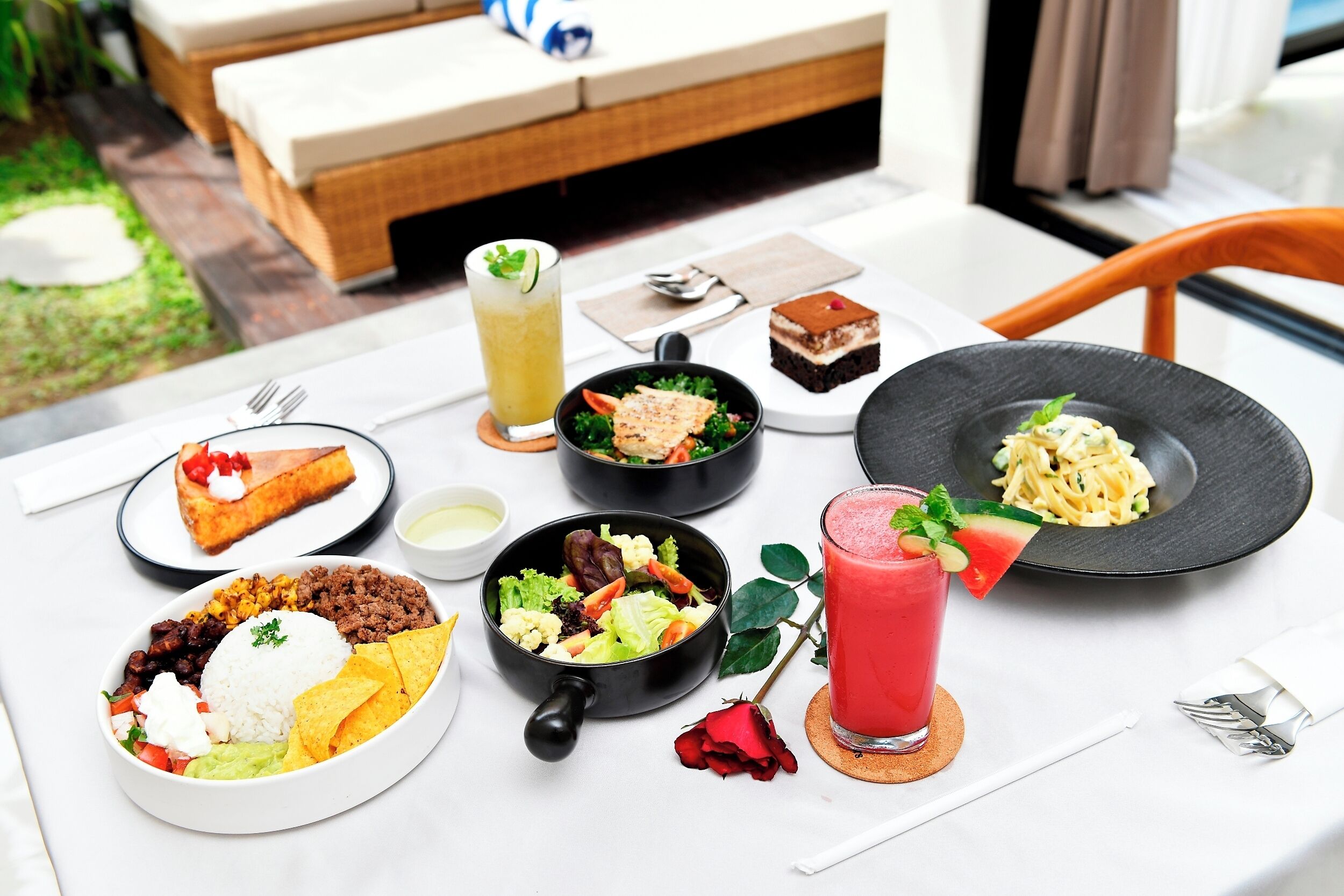 A table set with various dishes including rice, beans, salad, and desserts, with a rose and a drink on the table.