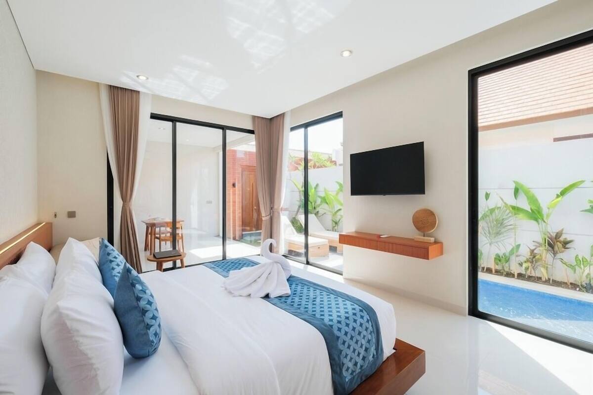 Ein modernes Hotelzimmer mit einem großen Bett, einem Flachbildfernseher, einem kleinen Tisch und Blick auf einen Swimmingpool.