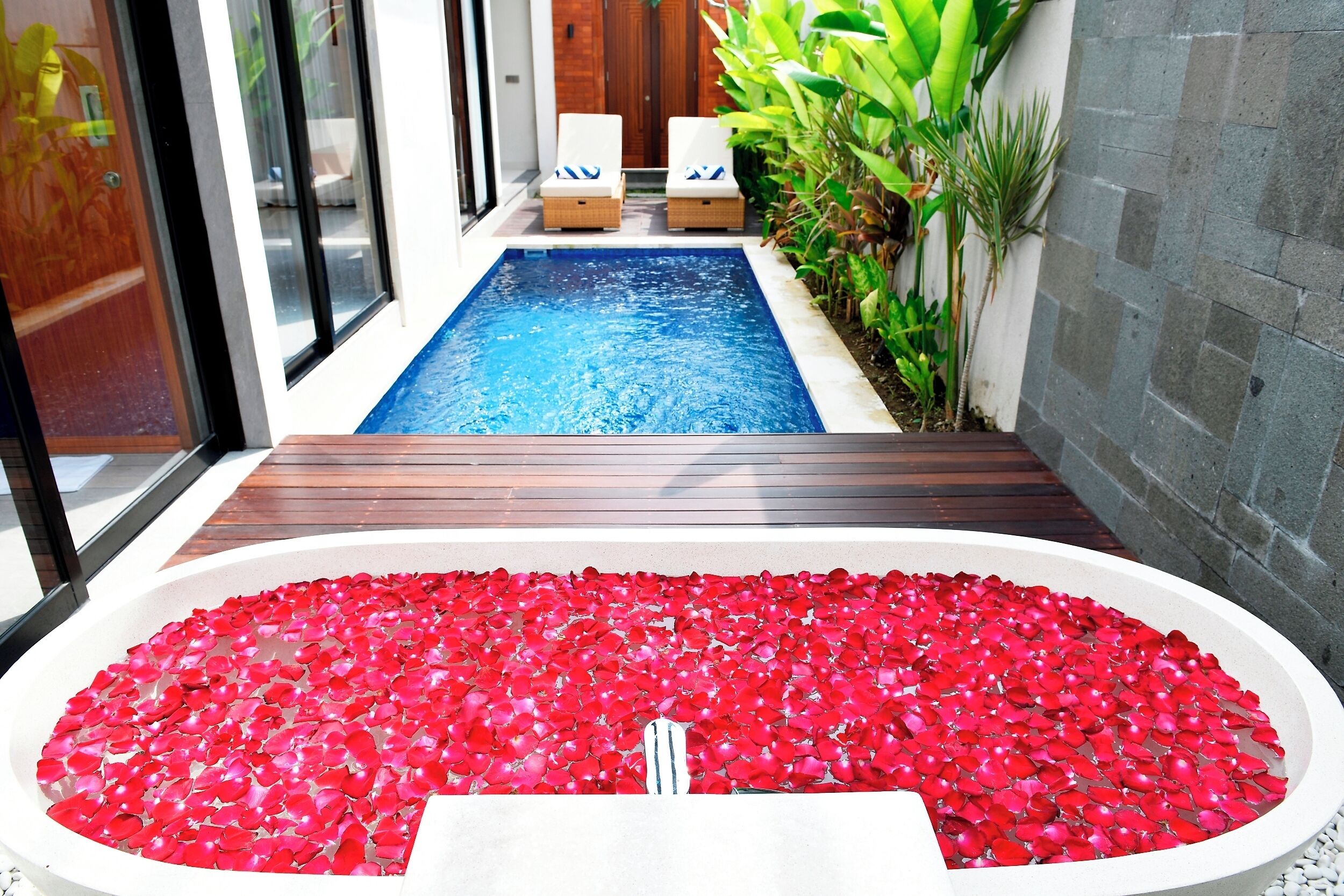 A swimming pool with a wooden deck, a jacuzzi with red petals, and lounge chairs.