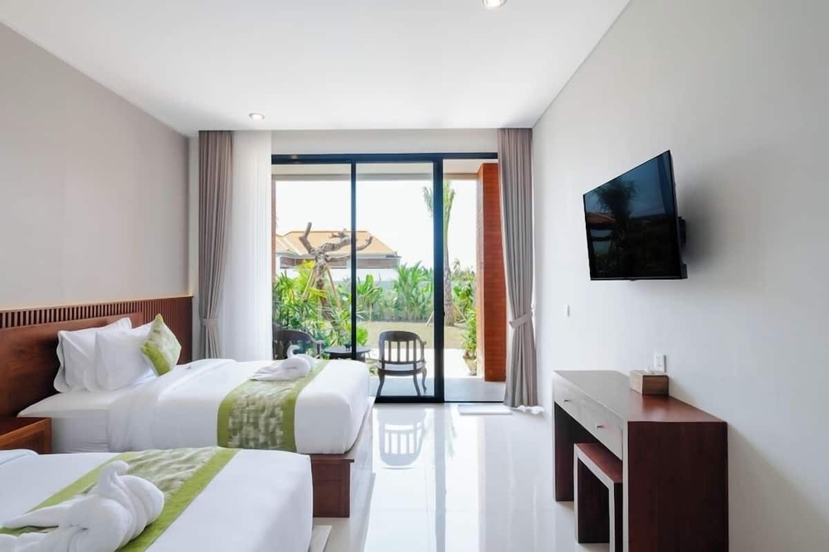 Ein Hotelzimmer mit zwei Betten, einem an der Wand montierten Fernseher und einem Balkon mit Blick auf Grünflächen und ein Gebäude.