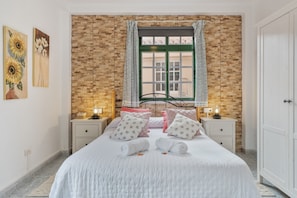 A bedroom with a brick wall, a window with green shutters, a bed with white bedding, two bedside tables with lamps, and artwork on the wall.