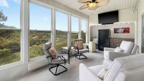 A spacious room with large windows offering a view of a forested area. There are two armchairs and a sofa, a ceiling fan, and a flat-screen TV mounted on the wall. - Vacay Getaway - Condo for 8 (Branson)