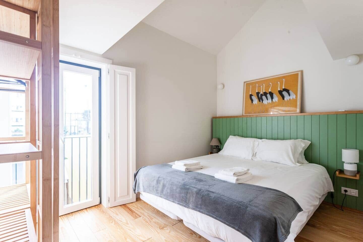 Une chambre à coucher avec un lit, une tête de lit verte, un tableau, une table de chevet et une fenêtre donnant sur l’extérieur.
