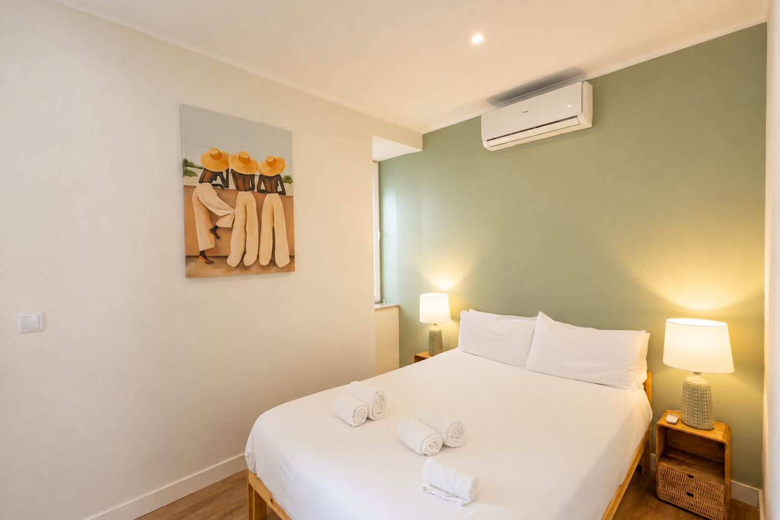 A hotel room with a bed, two bedside lamps, an air conditioning unit, and a wall-mounted artwork.