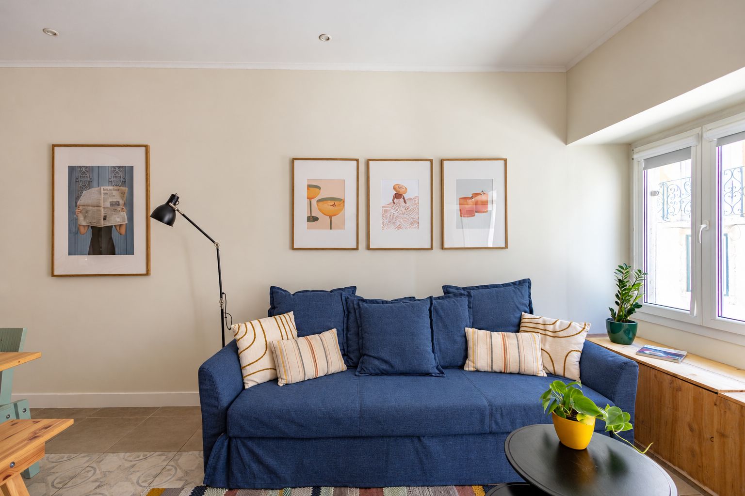 A living room with a blue sofa, framed artwork on the wall, a floor lamp, and a window with a view.