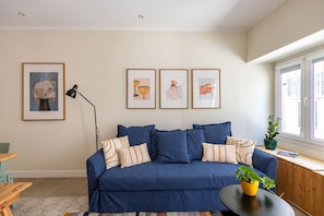 A living room with a blue sofa, framed artwork on the wall, a floor lamp, and a window with a view. - Stylish Oasis in Trendy Historic Centre Quarter (Lisbon)