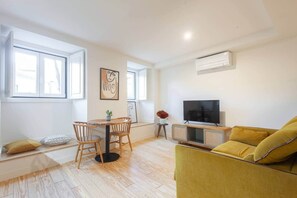 A modern living room with a yellow sofa, a TV on a wooden stand, a dining table with chairs, and artwork on the walls. - Modern and Quiet Central Studio Retreat (Lisbon)