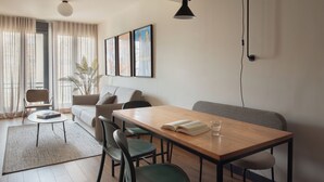 A modern living room with a wooden dining table, green chairs, a sofa, and a coffee table. There are framed pictures on the wall, a potted plant, and a lamp. - Líbere Madrid Malasaña (Madrid)