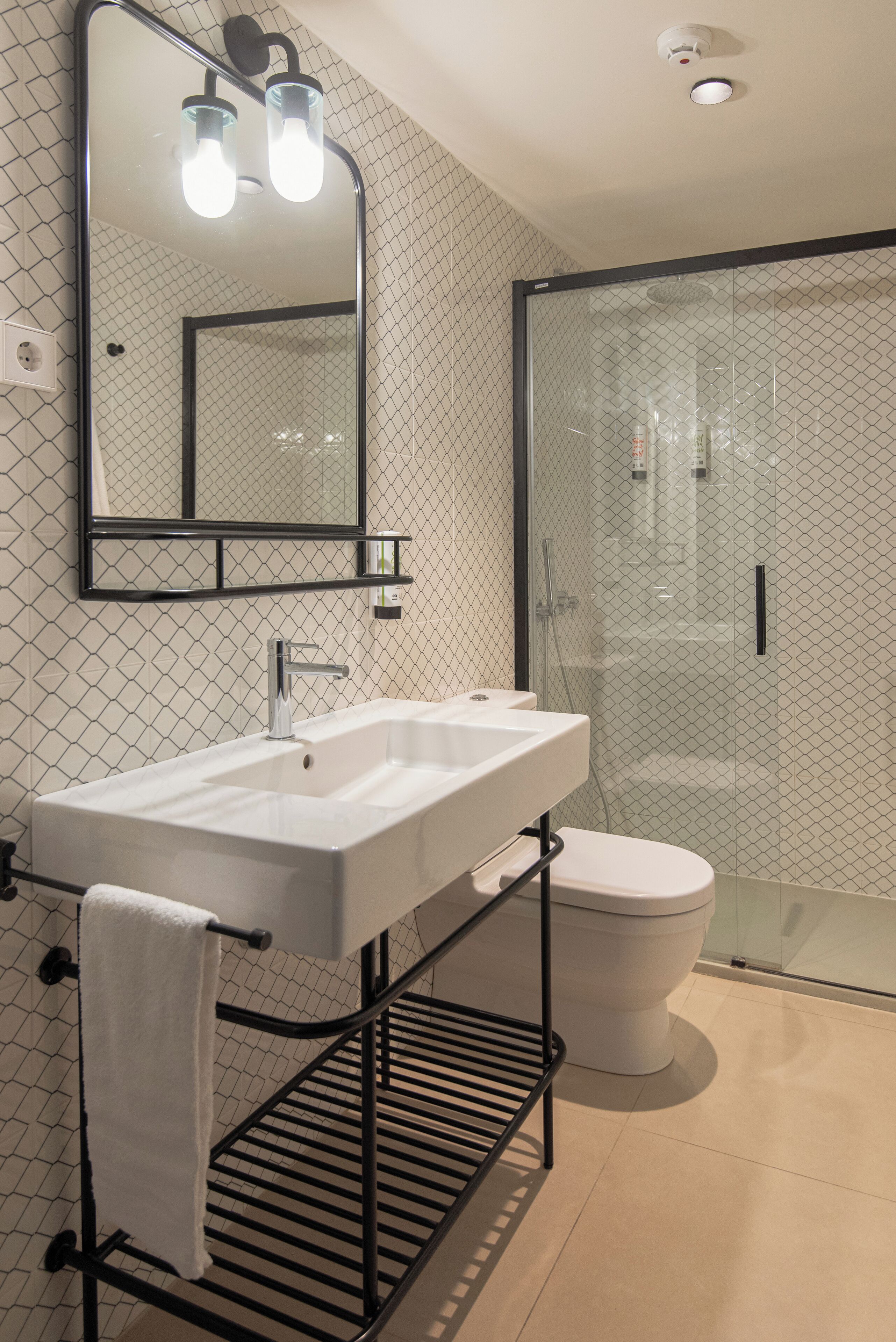 Un baño moderno con lavamanos, espejo y inodoro blancos, con cabina de ducha de cristal y un perchero metálico.