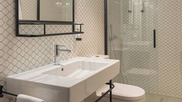 Un baño moderno con lavamanos, espejo y inodoro blancos, con cabina de ducha de cristal y un perchero metálico.
