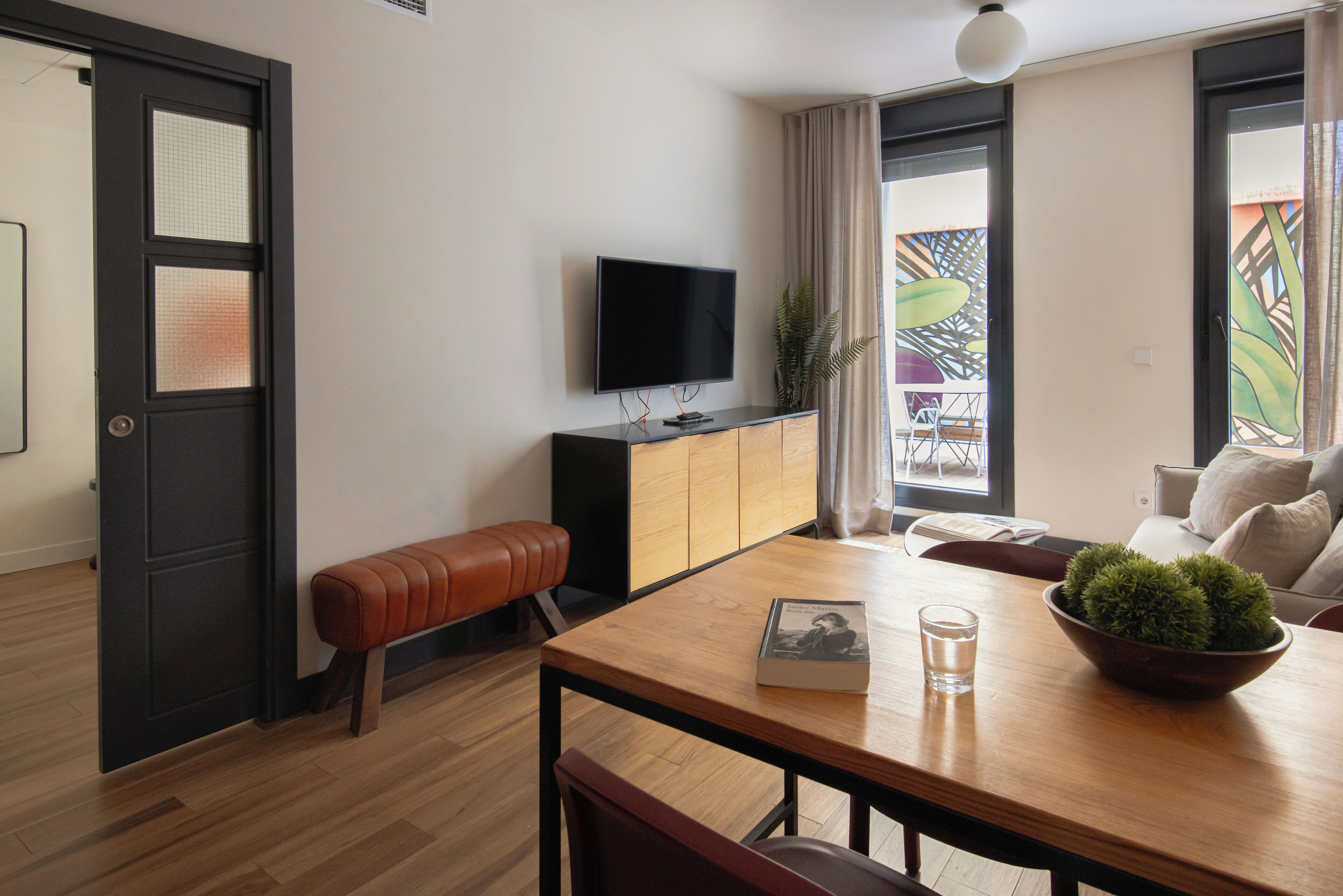 A modern living room with a wooden dining table, a bench, a television mounted on the wall, and a window with curtains.