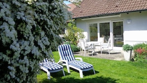 A house with a tiled roof, a patio with two striped lounge chairs, and a large flowering bush. - Apartment Noack, 50 m² with Private Terrace, Garden Area, and Wi-Fi (Immenstaad am Bodensee)