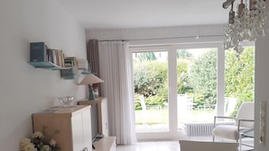A modern living room with a glass table, a sofa, a bookshelf, and a view of a garden through large windows. - Apartment Noack, 50 m² with Private Terrace, Garden Area, and Wi-Fi (Immenstaad am Bodensee)