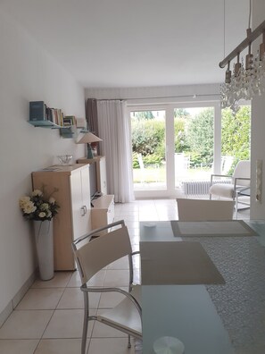 A modern living room with a glass table, a sofa, a bookshelf, and a view of a garden through large windows. - Apartment, 50 sqm, with private terrace, garden area, and Wi-Fi (Immenstaad am Bodensee)
