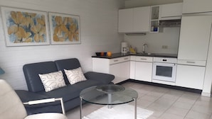 A modern living room with a sofa, a glass coffee table, and a kitchen area with white cabinets and appliances. - Apartment Noack, 50 m² with Private Terrace, Garden Area, and Wi-Fi (Immenstaad am Bodensee)
